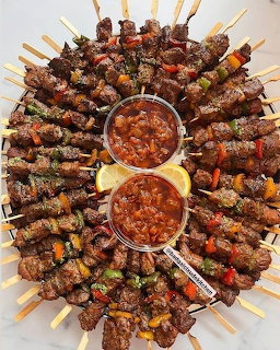 Circular platter of grilled meat skewers arranged around two bowls of red dipping sauce with lemon slices