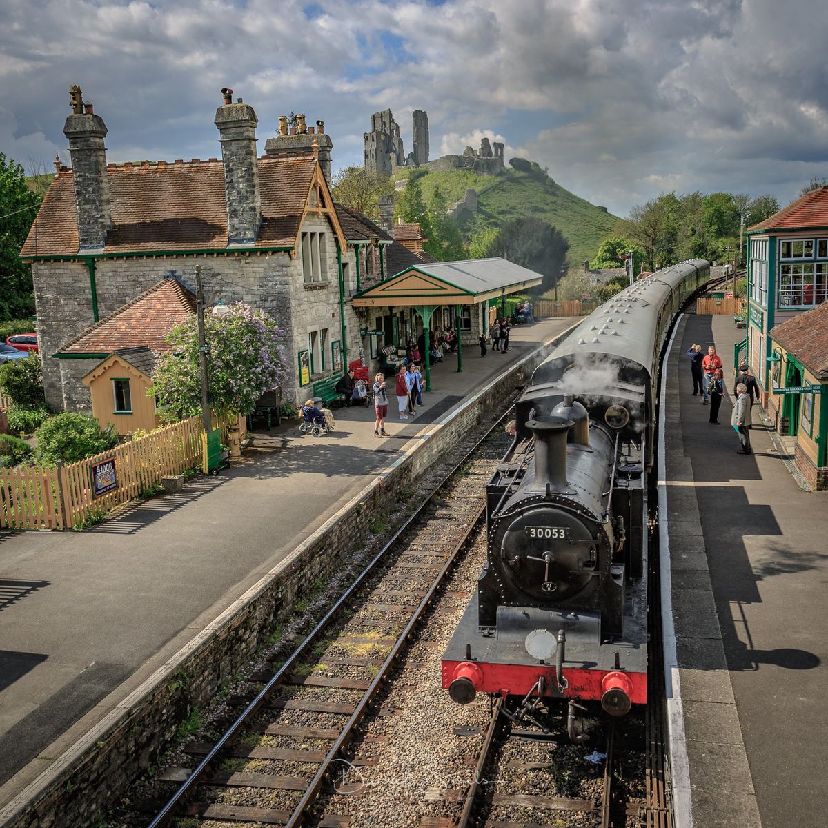 Black steam train at village station platform with castle on hill and cloudy sky