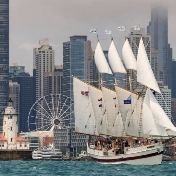 White-sailed tall ship in a harbor with city skyline, ferris wheel, and lighthouse