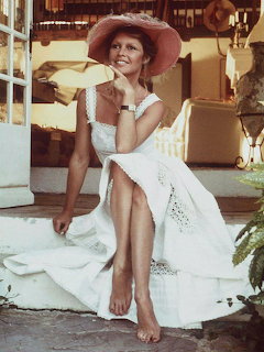 Woman in white summer dress and pink wide-brimmed hat sitting barefoot on a porch step