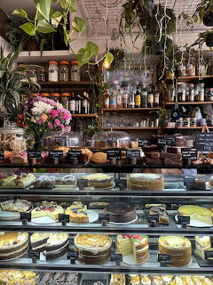 Cafe counter with glass display case full of cakes and desserts, shelves with jars, and hanging plants