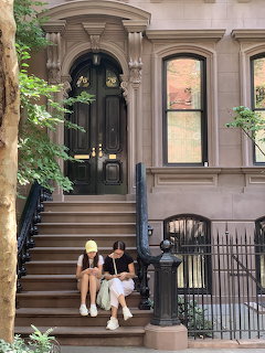 Two women sitting on the steps of a brownstone townhouse with black railings and trees
