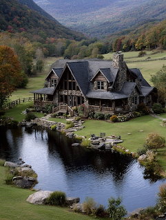 Rustic log house with dark roof beside a pond surrounded by green lawns and forested mountains