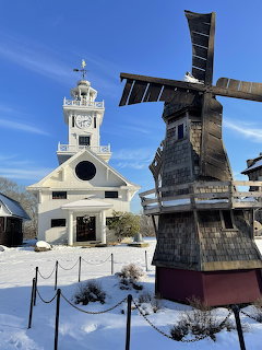 White church building with clock tower beside tall wooden windmill structure in snowy park setting