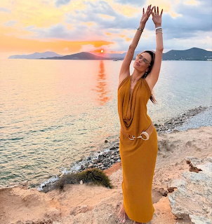 Woman in yellow dress with raised arms on beach at sunset with ocean and mountains behind