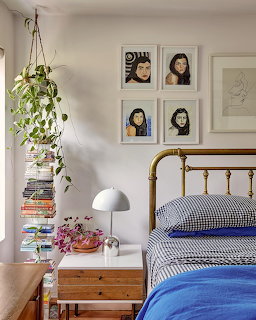 Cozy bedroom with brass bed, blue beding, nightstand, stacked books, hanging plant, and framed portraits