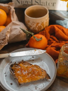 Toast with orange marmalade on a white plate next to fresh oranges, a marmalade jar, and a ceramic mug