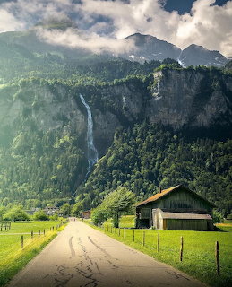 Country road leading to mountains with waterfall and wooden barn in a green alpine valley