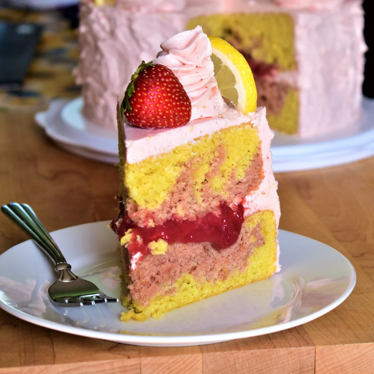 Slice of strawberry lemonade layer cake with pink frosting, fresh strawberry and lemon twist on white plate