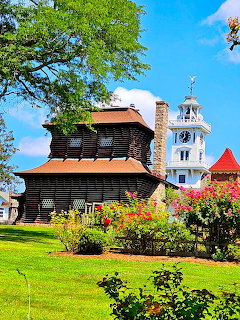 Dark wooden historic building with white clock tower and colorful rose garden on green lawn under blue sky