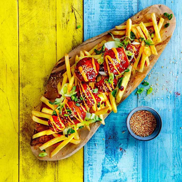 Loaded fries with fried bites, sauces, sesame seeds, and green onions on a wooden serving board