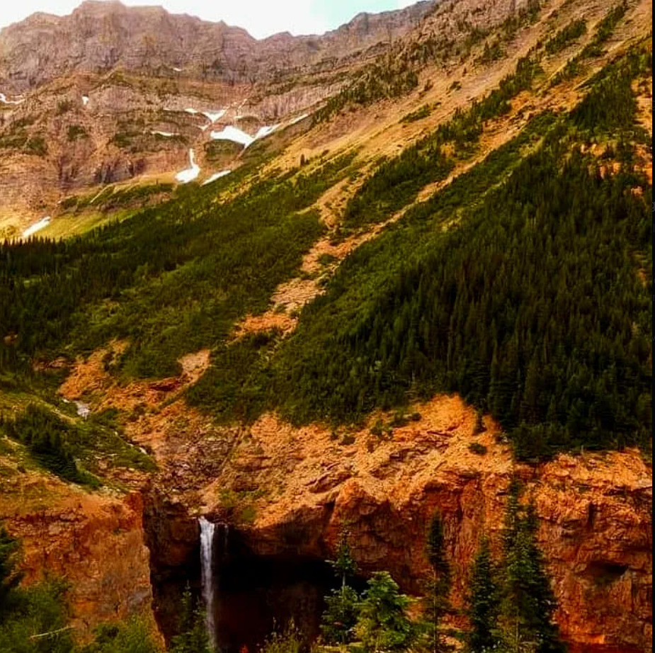Waterfall flowing from red rock cliffs with forested mountain slopes and rocky peaks in background