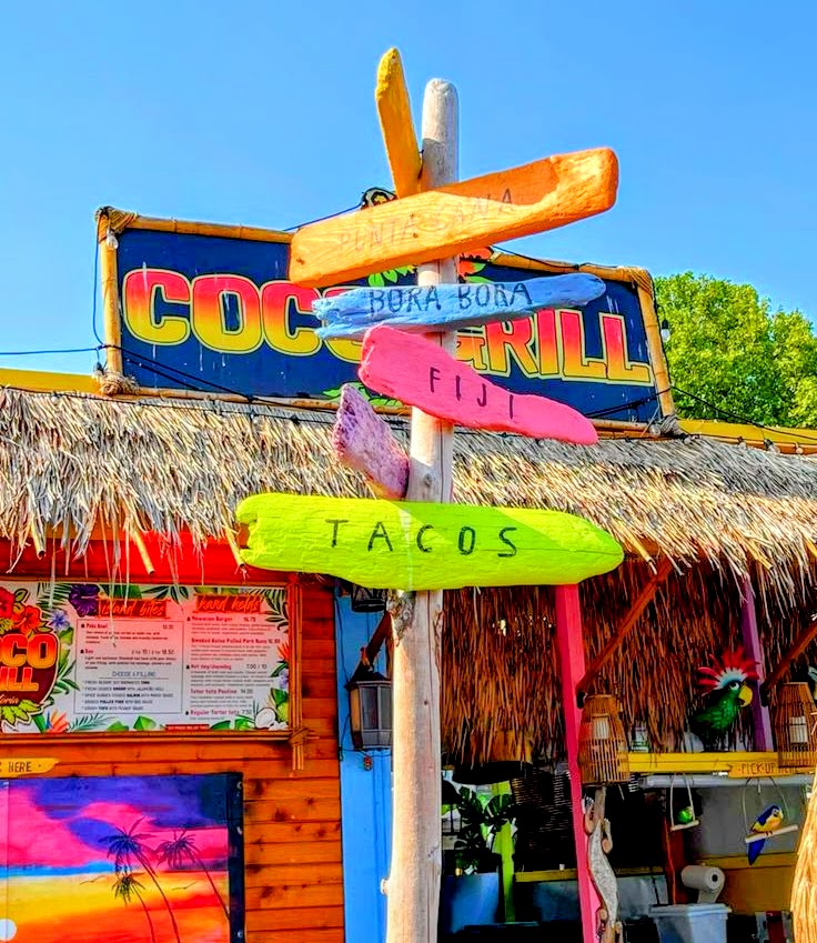 Colorful wooden signs reading Tacos, Fiji, and Hora Bora outside a tropical thatched bar called Coco Grill.