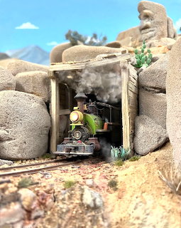 Model steam train emerging from a stone tunnel with smoke, surrounded by large boulders and desert plants