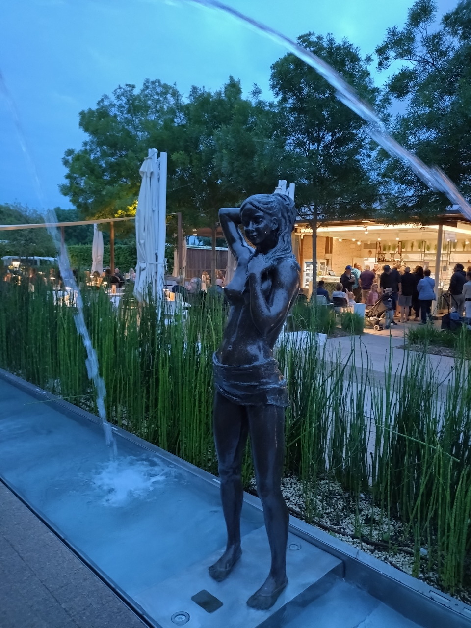 Bronze statue of a standing figure next to a water fountain in an outdoor plaza at dusk