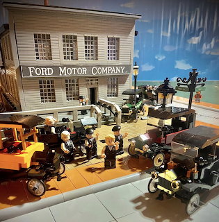 Lego model of Greenfield Village with Ford Motor Company building, vintage black cars, and minifigures