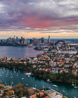 Aerial view of Sydney harbor at sunset with skyline, boats, and residential neighborhoods under colorful clouds