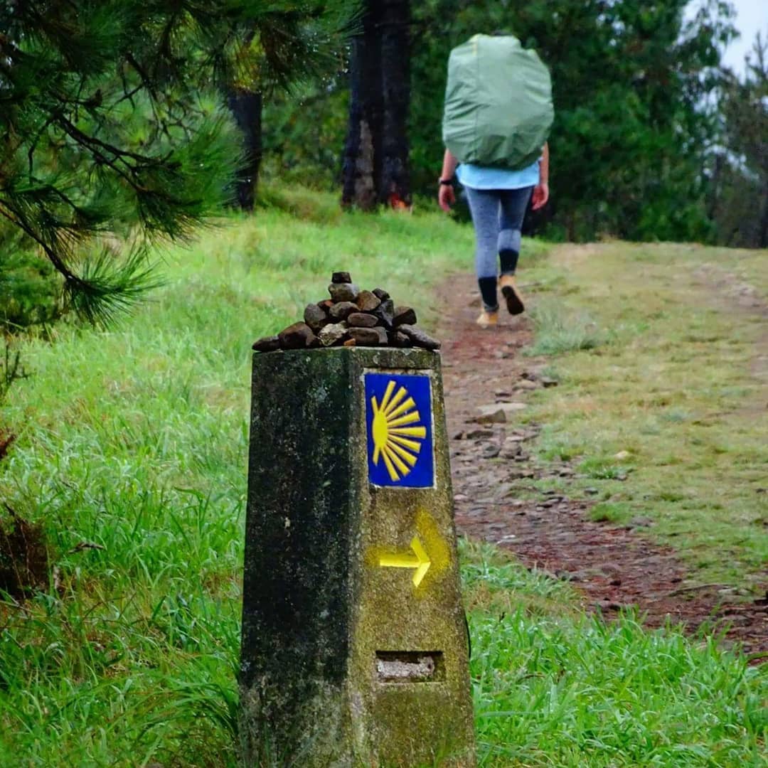 Stone marker with yellow scallop shell on Camino de Santiago trail, hiker walking ahead on grassy path