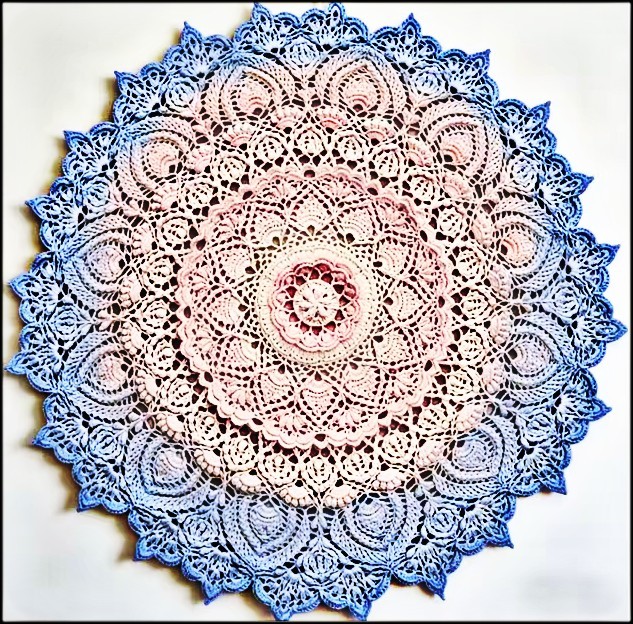 Circular crochet mandala doily with pink center fading to blue edges, intricate lace pattern on white background