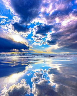 Stormy clouds reflected in wet sand on a beach with blue sky and ocean in the distance
