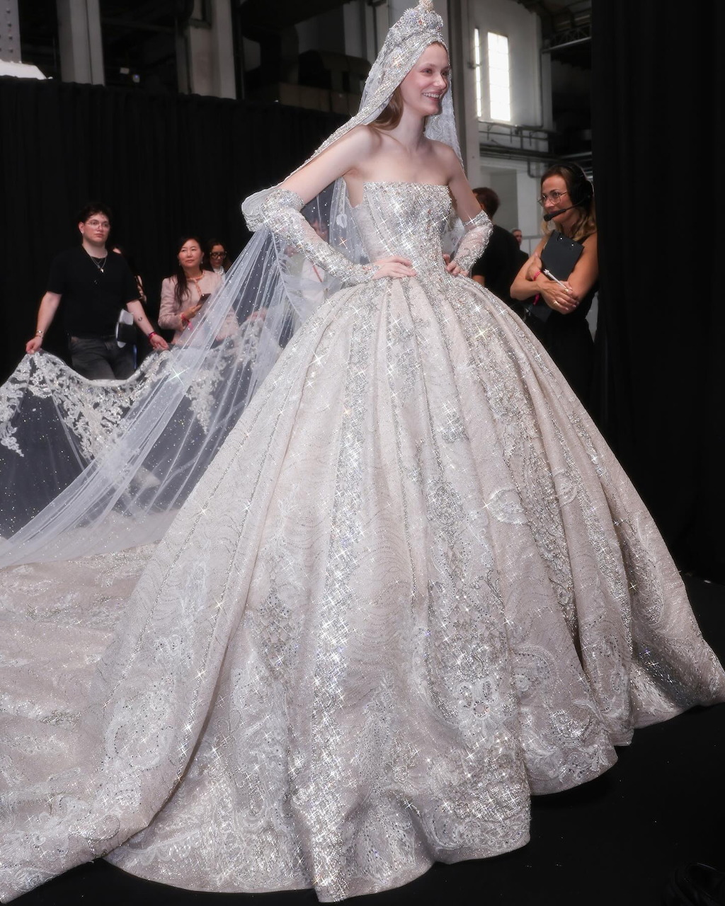 Model in elaborate silver beaded ball gown at bridal fashion show with assistants adjusting the dress