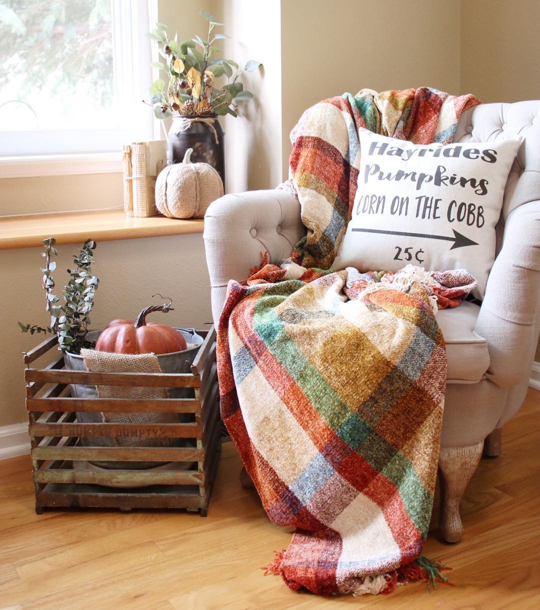 Cozy armchair with plaid throw, harvest pillow, pumpkins, and rustic crate near a window