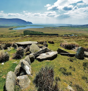 Stone ruins in grassy foreground overlooking fields, water, and distant hills beneath a cloudy sky