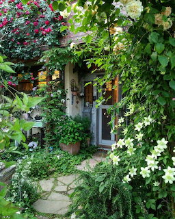 Cottage entrance with a stone path, dense greenery, and blooming white and pink flowers