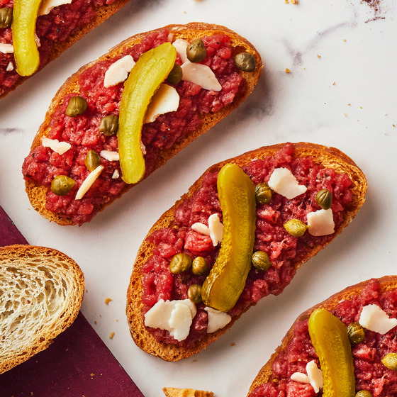 Three crostini with steak tartare, pickle slices, and white cheese on marble counter