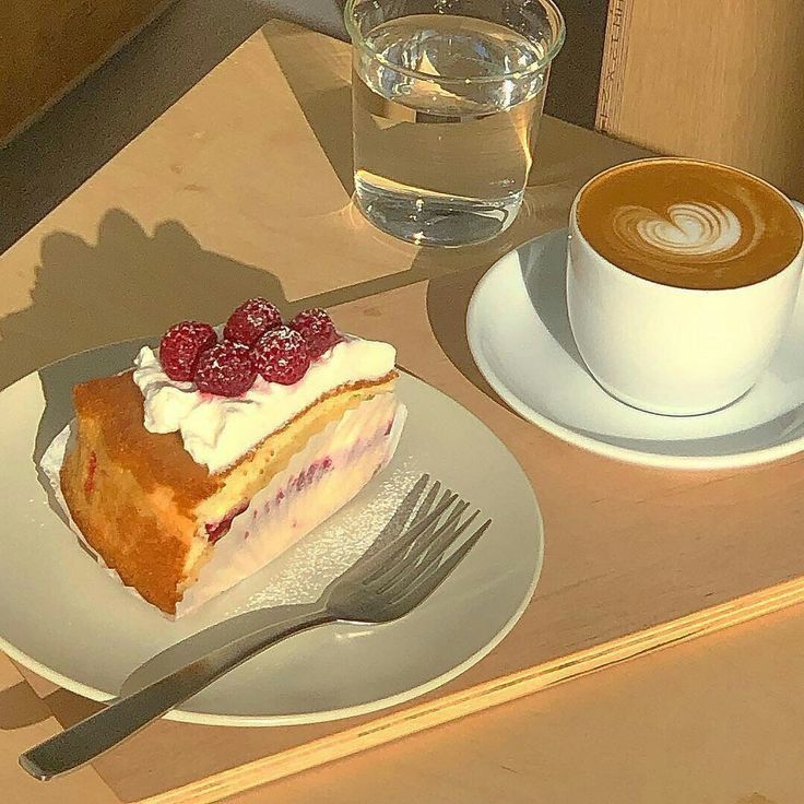 Slice of raspberry cream cake with a latte, glass of water, and fork on a sunlit table