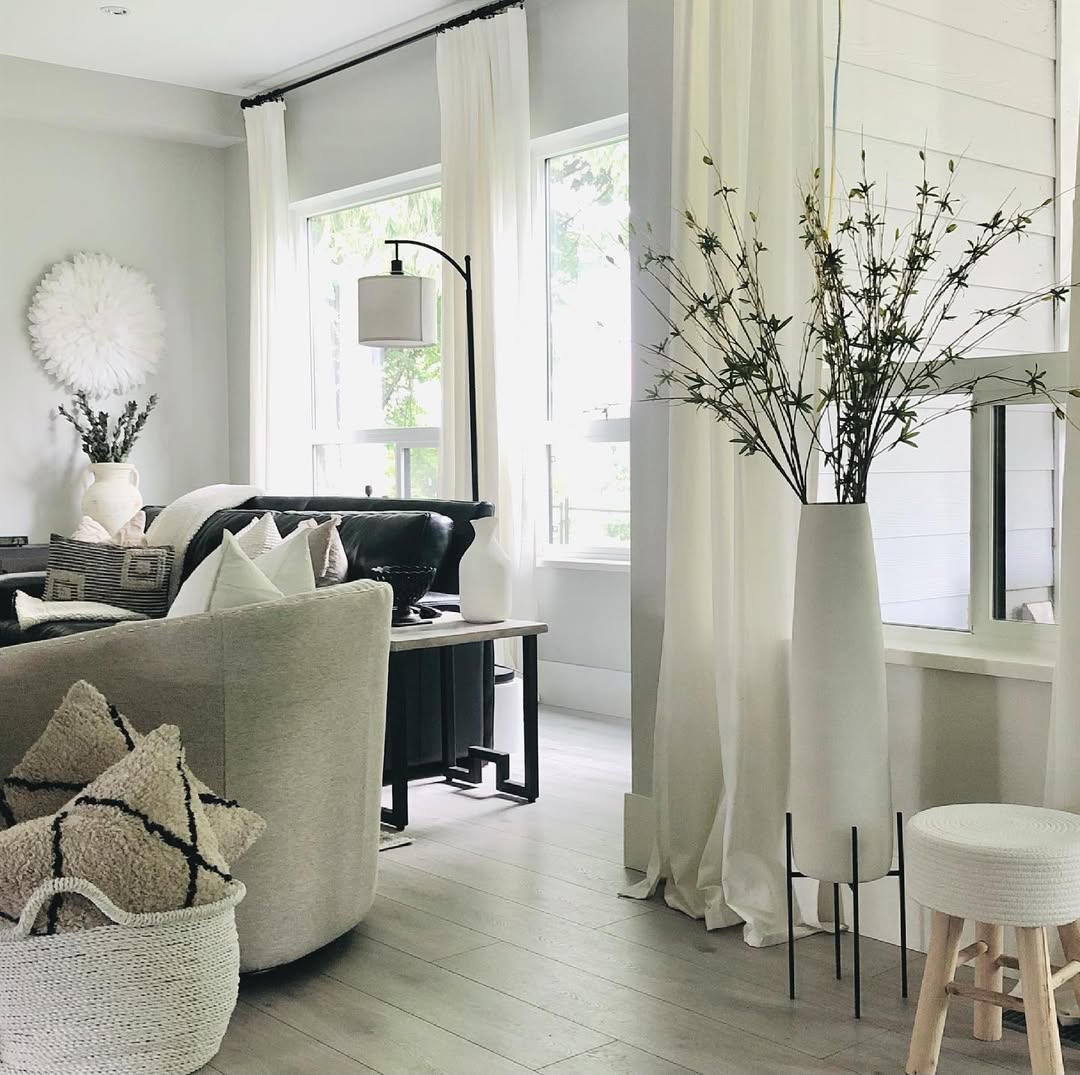 Bright farmhouse living room with white walls, cushioned chairs, sheer curtains, and a vase of branches