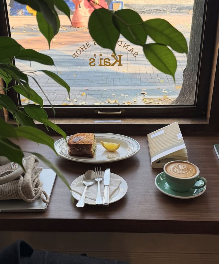 Cafe table by window with latte art in green cup, cake slice, cutlery, and indoor plant