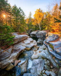 Rocky stream flowing between large boulders with autumn trees and sunburst in a forest