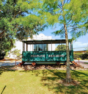Old green railroad passenger car labeled People's Railroad sitting outdoors under trees