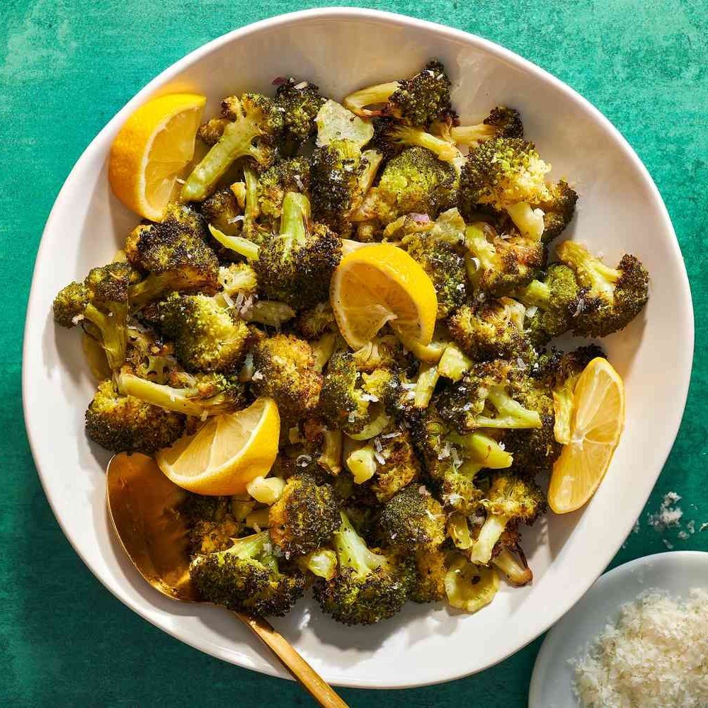 White plate of roasted broccoli with lemon wedges and grated cheese on a teal background