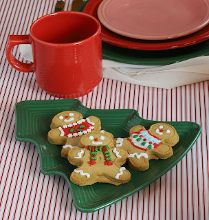 Two gingerbread cookies decorated with icing on a green leaf-shaped plate with red mug and colorful plates