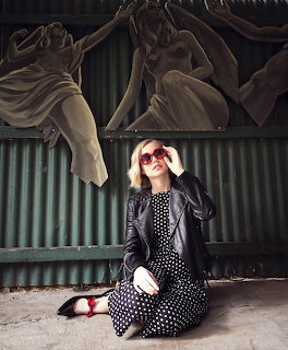 Blonde woman in polka dot dress and leather jacket sitting in front of painted corrugated wall