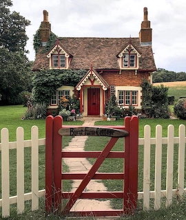 Brick cottage with red door and gate, white picket fence, green lawn and trees