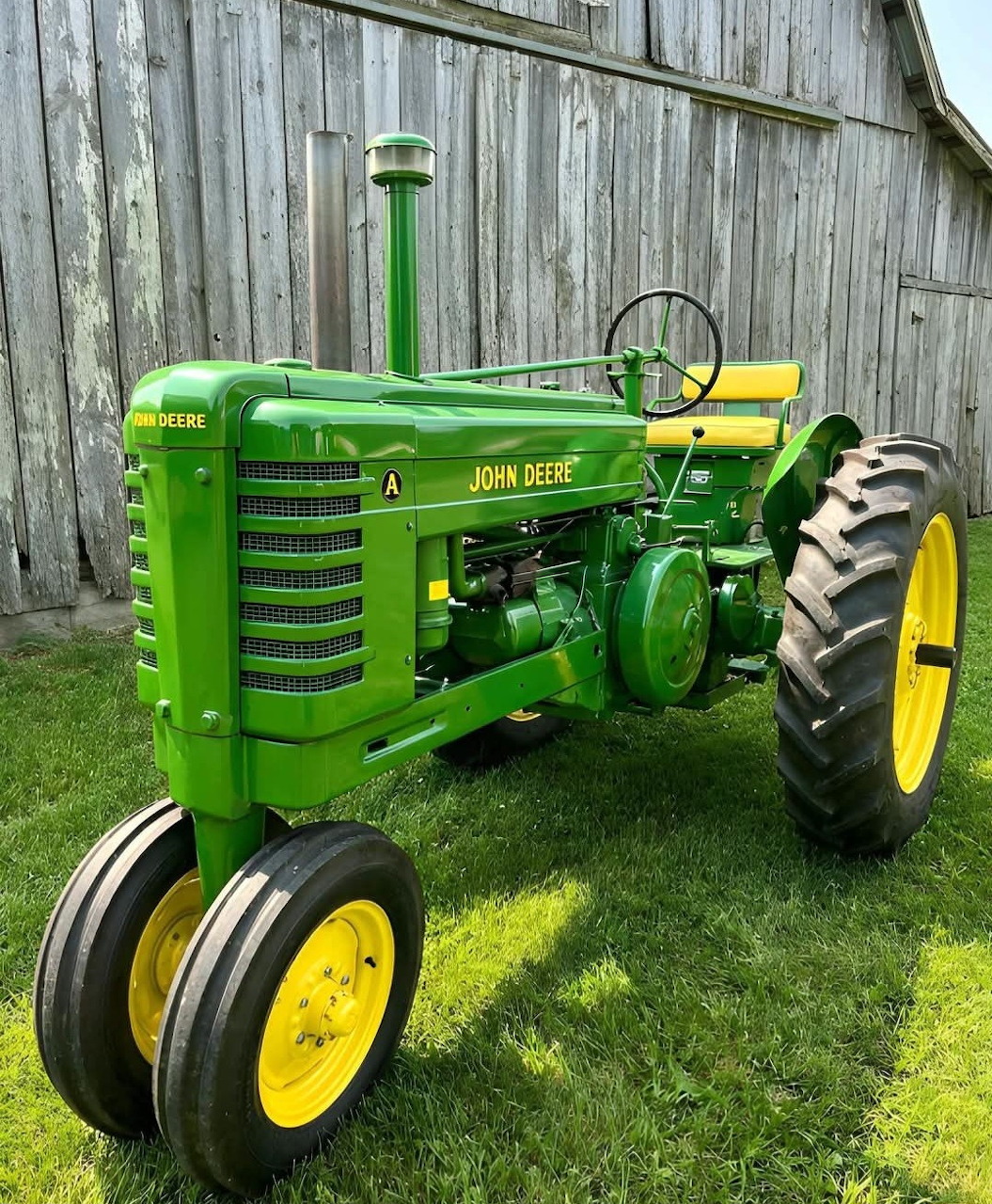 Green John Deere Model A tractor on grass beside a weathered wooden barn