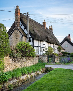 White thatched cottage with timber framing and stone wall along a village lane