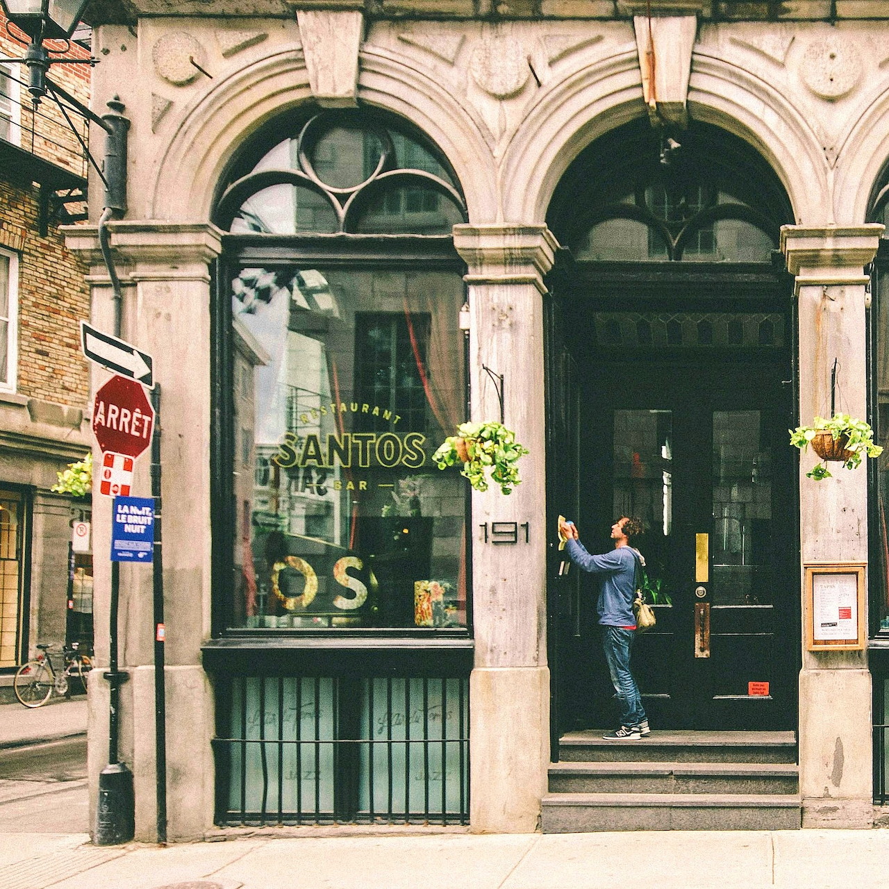 Stone building facade in Old Montreal with arched doors, Santos storefront, hanging plants, and a person photographing