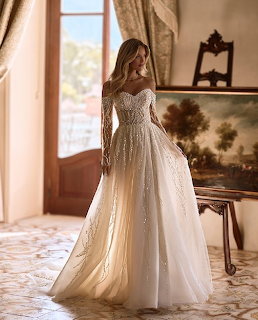Woman in a sparkling off-shoulder bridal gown with long lace sleeves standing in a sunlit room
