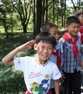 Smiling boy in Barney t-shirt saluting outdoors with children in background among trees
