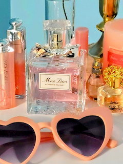 Pink heart-shaped sunglasses in front of various perfume bottles on a pink surface with turquoise background
