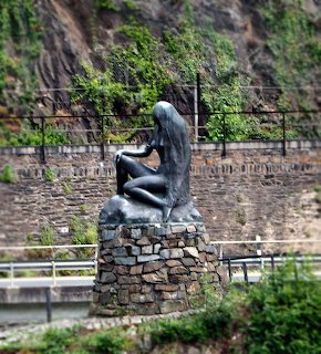 Seated statue on a stone pedestal beside a rocky wall with greenery