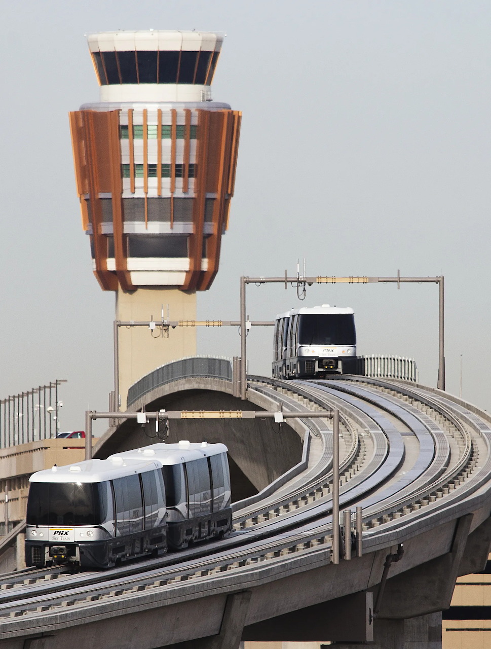 Two white automated trains on curved elevated tracks beside an airport control tower