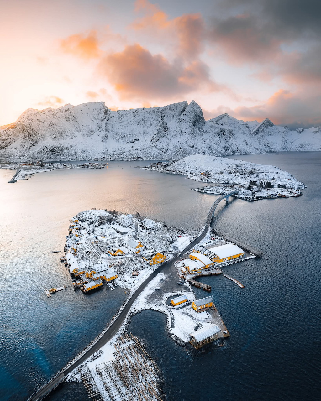 Aerial view of snowy Lofoten Islands fishing village surrounded by fjord and mountains at sunset, Norway
