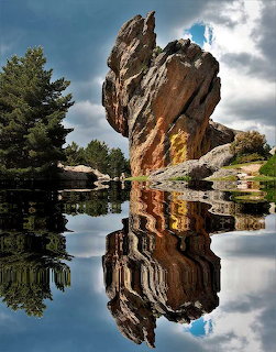 Tall rock formation mirrored in calm water with trees and cloudy sky