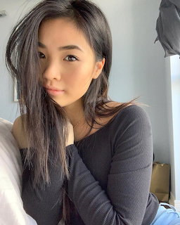 Young Asian woman with long dark hair taking a selfie in a gray long-sleeve top indoors