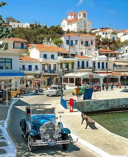 Greek island waterfront with white houses, church on hill, vintage car, and harbor with boats